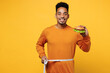Young cheerful man wear orange sweatshirt casual clothes eat burger hold measure tape on waist isolated on plain yellow background studio. Proper nutrition healthy fast food unhealthy choice concept.