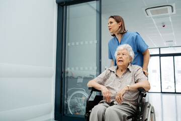 Portrait of nurse pushing senior patient in wheelchair across hospital corridor. Emotional support for elderly woman.