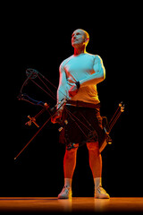 Wall Mural - Full-length image of man, archer standing with archery bow and arrows against black studio background in neon light. Concept of professional sport and hobby, competition, action, game