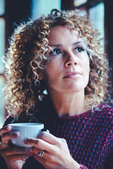 Wall Mural - Portrait of thoughtful attractive adult woman at home holding a cup of tea and looking outside the windows - Pretty curly young mature female people enjoy relax indoor in cozy cabin