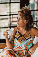 Wall Mural - Elegant attractive young adult woman with warm knitted wool colored dress sitting at home drinking from cup and looking outside - concept of trendy beautiful female people enjoying leisure relax 