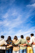 © Xavier Lorenzo - Vertical photo. Rear view of young group of happy multiracial friends hugging each other standing in a row looking blue sky