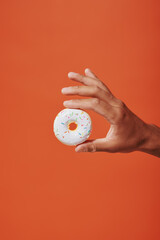 Wall Mural - cropped view of person holding glazed vanilla donut with sprinkles on orange background, white icing