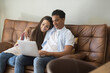 © simona - Happy young relaxed couple sitting on sofa with laptop on laps, discussing funny movie, enjoying watching comedian film, shopping online or web surfing, full length front view, leisure pastime concept