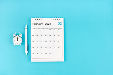 February 2024, Monthly desk calendar for 2024 year and pen with alarm clock on blue.