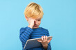 Young child holding digital tablet, watching videos, rubbing his eyes, isolated over pastel blue background. Children and technology, screen time control and eye health.