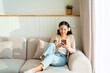 © Kittiphan - Happy young Asian woman with mobile phone listening music in headphones and relaxing on safa at home