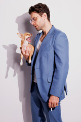 Fashionable man in blue suit and eyeglasses holding cigarette and chihuahua dog on white