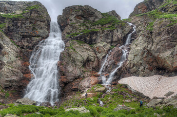 Wall Mural - Waterfall in a mountain valley. small figures of people against the backdrop of mountains.