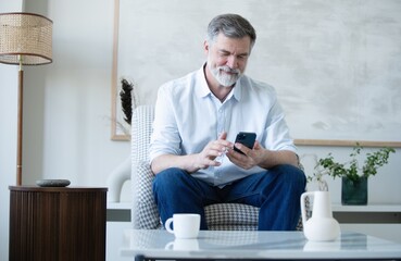 Wall Mural - Smiling mature freelance businessman using mobile phone checking social media network or message chat sitting on chair at home.