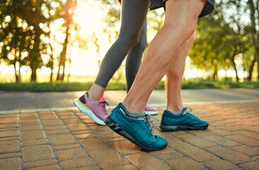 Wall Mural - Heathy and active lifestyle. Close up of male and female legs in blue and pink sneakers jogging on cobblestones path at summer park. Caucasian aged couple spending morning time for outdoors activity.