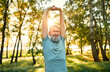 © HBS - Active and healthy retirement. Portrait of elderly man stretching arms and back before morning exercises on fresh air. Caucasian person warming up while preparing for cardio workout at summer park.