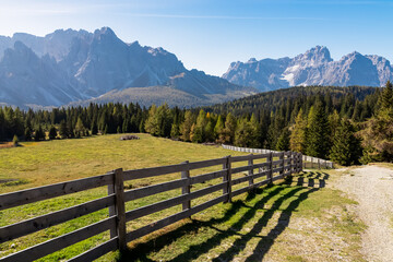 Scenic hiking trail along wooden fence on alpine meadow on Nemes Alm (Rifugio Malga Nemes) in Carnic Alps, South Tyrol, Italy. View of massive mountain ridges of majestic untamed Sexten Dolomites