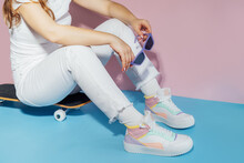 No face female in white outfit and retro style high-top multicolor sport sneakers shoes with sunglasses sitting on skateboard on blue pink background. Vintage retro fashion style of 80s - 90s vibes