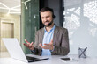 © Liubomir - Smiling young man sitting at desk in office wearing headset and talking gesturing hands on video call via laptop.