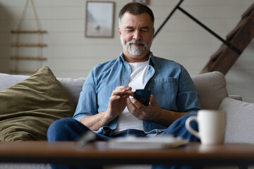 Sticker - Happy smiling senior man using smartphone device while sitting on sofa at home