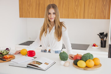 Portrait of a cute blonde girl smiling and reading a recipe book in the kitchen on the table among ingredients for cooking.