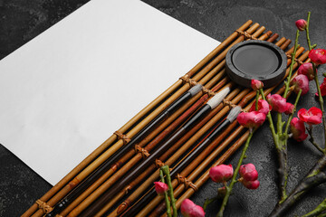 Wall Mural - Composition with blank sheet of paper, bamboo mat, brushes, ink and blooming branch on dark background, closeup. International Haiku Poetry Day