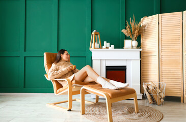 Poster - Young woman with cup of tea resting in armchair near fireplace at home