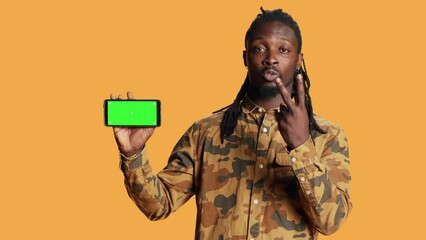 Wall Mural - Cheerful adult holds smartphone with greenscreen in studio, presenting isolated copyspace display on mobile phone layout. Young person showing blank chromakey mockup on device.