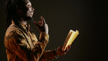 Wall Mural - Young adult reading literature book on camera, standing over black background and enjoying lecture as hobby. African american person having fun with fiction or action novel story. Handheld shot.