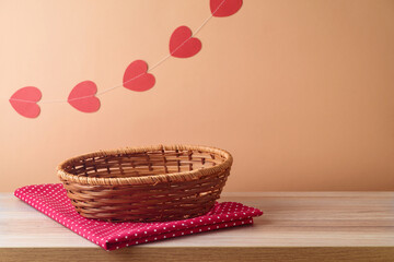 Wall Mural - Empty basket on wooden table with tablecloth and heart shape garland over peach color background.  Valentines day mock up for design and product display