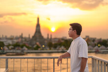 Tourist Man Enjoys View To Wat Arun Temple In Sunset, Traveler Visits Temple Of Dawn Near Chao Phraya River From Rooftop Bar. Landmark And Travel Destination In Bangkok, Thailand And Southeast Asia