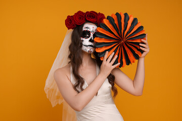 Wall Mural - Young woman in scary bride costume with sugar skull makeup, flower crown and paper decoration on orange background. Halloween celebration