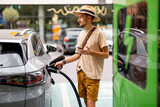 Fototapeta Sypialnia - Man in hat plugs a cable in electric vehicle, while standing with phone on a public charging station outdoors. Concept of travel by electric car and green energy for driving