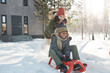 © pressmaster - Cheerful young woman in warm winterwear pushing sledge with her cute son during winter stroll along snow path against country house