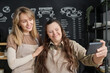 © pressmaster - Young smiling waitress with mental disability holding smartphone in front of herself while taking selfie with happy blond colleague in cafe