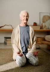 Wall Mural - Active senior woman with closed eyes sitting on knees in front of camera and keeping balance during relaxation yoga exercise at home