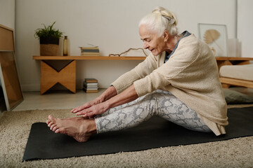 Wall Mural - Side view of aged active woman in casualwear stretching legs and arms forwards while sitting on the floor in front of camera at home