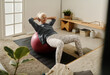 © pressmaster - Senior active woman doing sit ups on fitball during morning physical training on mat on the floor of bedroom with wooden furniture