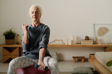 Wall Mural - Aged woman in activewear sitting on fitball and practicing physical exercise for arm muscles with dumbbells in front of camera