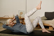Active female pensioner in sportswear lying on mat in bedroom and doing physical exercise with one leg raised and hands on back of head