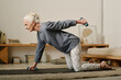 Side view of senior active woman in leggins and pullover standing on knees on the floor of bedroom and doing exercise with dumbbells