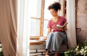 Wall Mural - Young smiling pretty mixed race girl sits on windowsill with porcelain cup of tea and book