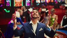 Portrait Of An Excited Young Man Catching Money That Is Flying From The Sky. Successful Gambler Won A Jackpot In A Casino. Concept Of Gambling, Betting, Finance, Luxury And Success. Slow Motion