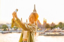 Young Asian Woman Traveler Taking A Selfie While Enjoying The Sunset Moments Of Wat Arun By The Chao Phraya Riverbank In Bangkok, Thailand