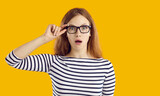 Fototapeta  - Young student girl very surprised by something. Beautiful nerdy young woman in striped top and glasses isolated on yellow background looking at camera with funny, shocked, astonished face expression