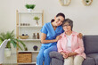 A beautiful, young, happy, smiling nurse kindly embraces her patient, an elderly, well-groomed, pretty gray-haired lady with glasses, sitting down on the edge of the sofa for photo posing for memory.