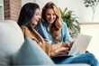 © nenetus - Two happy beautiful women using computer while planning their vacation sitting on the couch at home.
