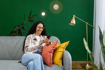 Beautiful Hispanic woman at home on sofa in the living room, the woman smiles contentedly holds the phone in her hands, uses applications on the smartphone, browses the Internet and social networks.