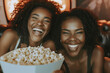 © MNStudio - Two cheerful female friends watching a tv film at home. Two black women eating popcorn with happy expressions on their faces. Having a movie night.