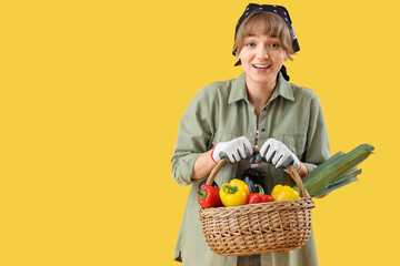 Wall Mural - Happy young female farmer with wicker basket full of different ripe vegetables on yellow background