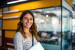 © apratim - Smiling young woman holding a laptop in a modern office environment, radiating confidence and positivity.