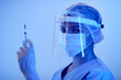 Medium close up shot of woman doctor in medical protective wear holding syringe while looking at camera in blue neon light