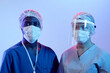 Medium close up shot of two diverse surgeons wearing medical protective wear looking at camera while standing at pastel blue background