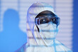 Medium close up shot of Black man scientist at blue background with shadow from window blinds wearing protective coverall and glasses while looking at camera